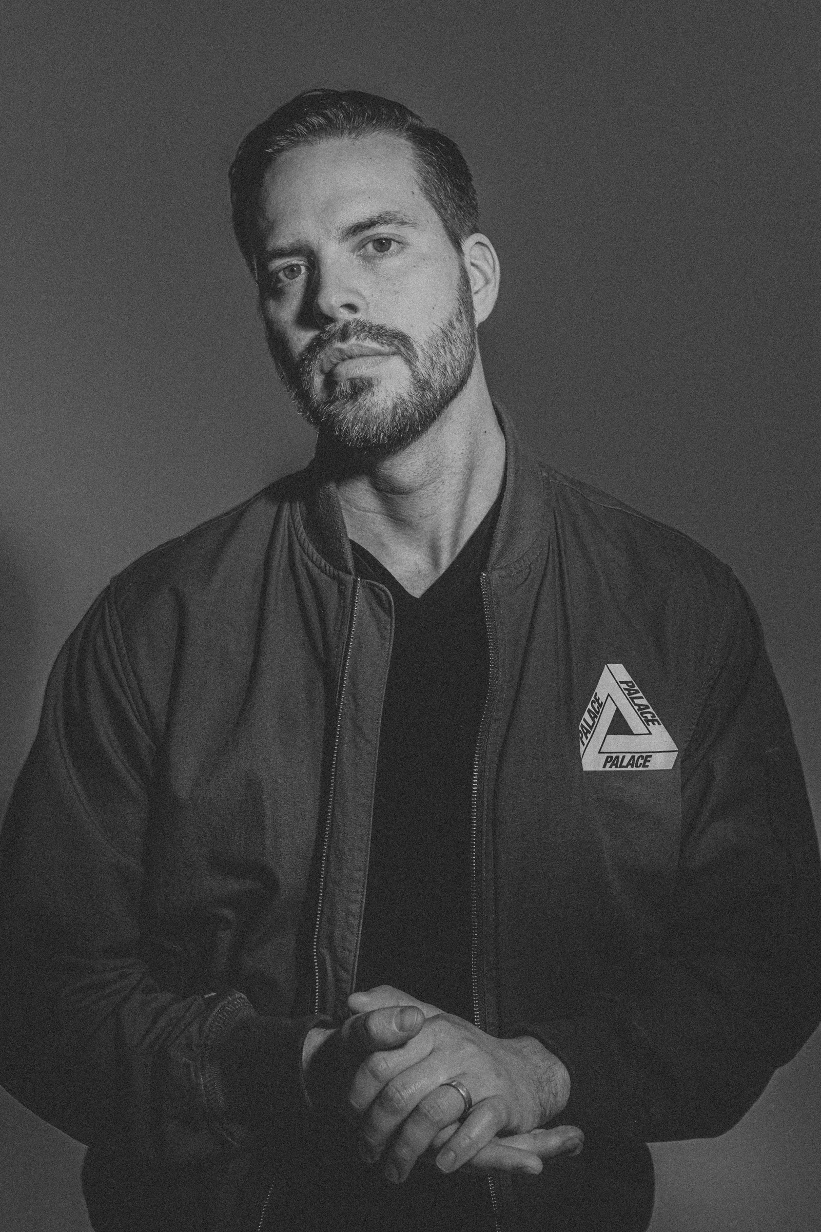 Black-and-white portrait of Patrick Dohan in a dark bomber jacket, hands clasped.